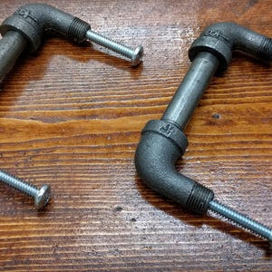 May include: Two industrial-style cabinet pulls made from black metal pipes and fittings. Each pull has a curved shape with a threaded end and a silver screw. The pulls are displayed on a wooden surface.