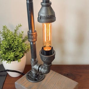 May include: Industrial-style desk lamp featuring a metal pipe design with a warm, glowing Edison bulb. The lamp is mounted on a rectangular wooden base. The lamp's height is approximately 12 inches.