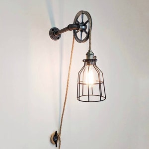 May include: A wall-mounted industrial-style light fixture with a pulley system and a caged light bulb. The fixture is made of metal and has a rustic finish. The light bulb is hanging from a rope that is threaded through the pulley.