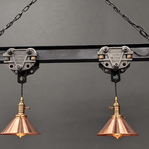 May include: A hanging light fixture featuring a dark gray metal I-beam suspended by chains. Two industrial-style metal trolleys are attached to the beam, each supporting a copper-toned conical pendant lamp with a visible light bulb at the base.