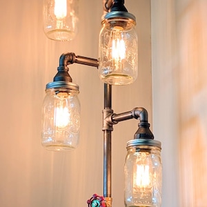 May include: A unique floor lamp crafted from metal pipes, featuring four clear glass mason jar shades. The lamp has a vintage industrial design with a red and green valve detail. The warm light from the bulbs creates a cozy ambiance.