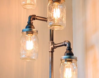 May include: A unique floor lamp crafted from metal pipes, featuring four clear glass mason jar shades. The lamp has a vintage industrial design with a red and green valve detail. The warm light from the bulbs creates a cozy ambiance.