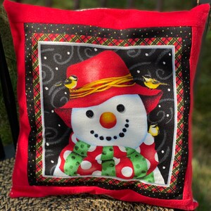 May include: Red throw pillow with a snowman wearing a red hat and green scarf with white polka dots. The snowman is surrounded by a black and white plaid border.