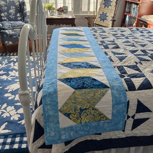 May include: A blue and yellow quilted bed runner with a chevron pattern. The runner is on a white bed with a blue and white quilt.