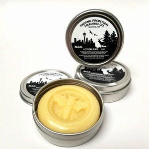 May include: A silver tin with a white label that reads "Fading Frontier Trading Co. Seattle, WA" and "Lotion Bar 1 oz". The tin contains a yellow lotion bar with a bee design.