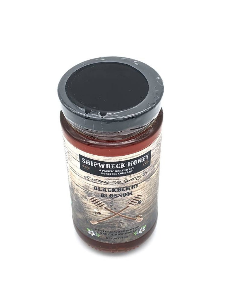 May include: A glass jar of Shipwreck Honey Blackberry Blossom honey. The jar has a black lid and a brown label with white text. The label says "Shipwreck Honey, A Pacific Northwest Honeybee Company, Blackberry Blossom, Western Washington, Unheated & Raw Honey, Net Wt 12 oz."