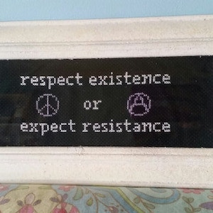 May include: White framed embroidery with a black background and white text that reads "respect existence or expect resistance". The embroidery includes a peace sign and a fist.