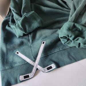 May include: A dark green hoodie with a white hockey stick patch on the front. The patch is a simple design of two crossed hockey sticks.
