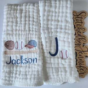 May include: Two white burp cloths with embroidered baseball designs. One cloth features a baseball, glove, bat, and hat with the name "Jackson" embroidered below. The other cloth features a baseball and the letter "J" embroidered in blue.