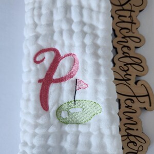 May include: White muslin baby burp cloth with pink embroidered letter P and a green embroidered golf ball with a pink flag.