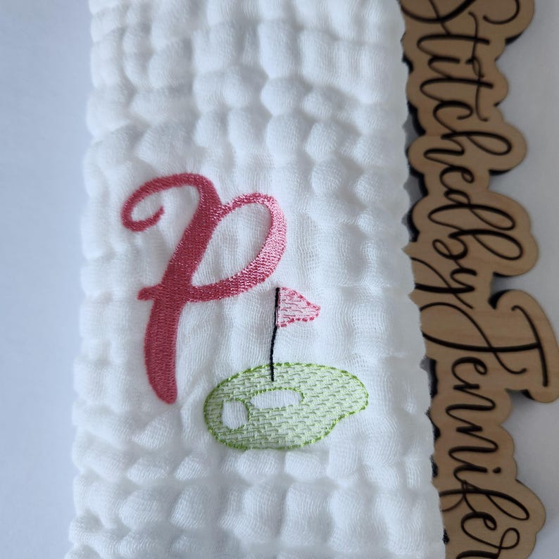 May include: White muslin baby burp cloth with pink embroidered letter P and a green embroidered golf ball with a pink flag.