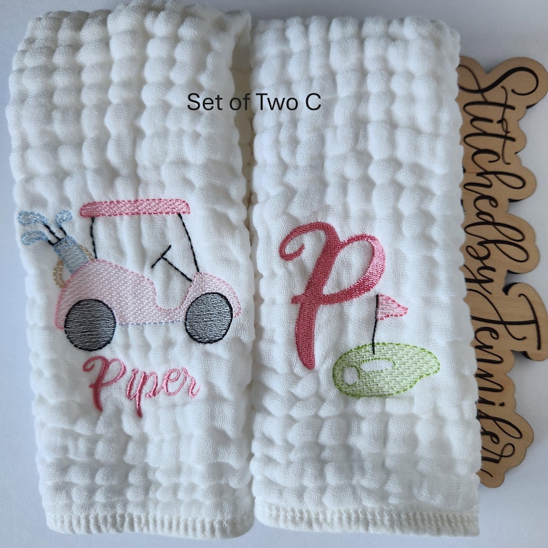 May include: Set of two white cotton burp cloths with embroidered designs. One cloth features a pink golf cart with the name "Piper" embroidered below. The other cloth has a pink letter "P" and a green golf ball with a flag.