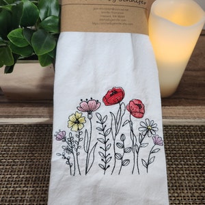 May include: White kitchen towel with embroidered flowers in shades of red, pink, yellow, and green. The embroidery features a variety of flowers and leaves, including poppies, daisies, and other wildflowers. The towel has a stitched border and the text "Stitched by Jennifer" is printed on the top.