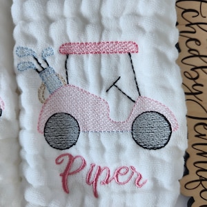 May include: A white muslin baby burp cloth with pink and black embroidery of a golf cart and the name "Piper".