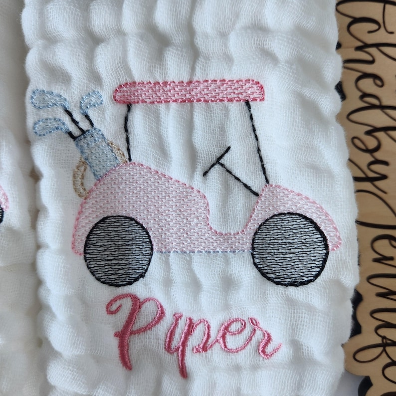 May include: A white muslin baby burp cloth with pink and black embroidery of a golf cart and the name "Piper".