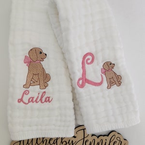 May include: Two white muslin burp cloths with pink embroidery. One cloth features a brown dog with a pink bow and the name "Laila" embroidered below. The other cloth features a pink letter "L" and a smaller brown dog with a pink bow.