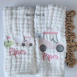 May include: Set of two white muslin burp cloths with pink and green embroidered golf cart designs and the name "Piper". The text "Set of Two A" is visible in the top right corner.
