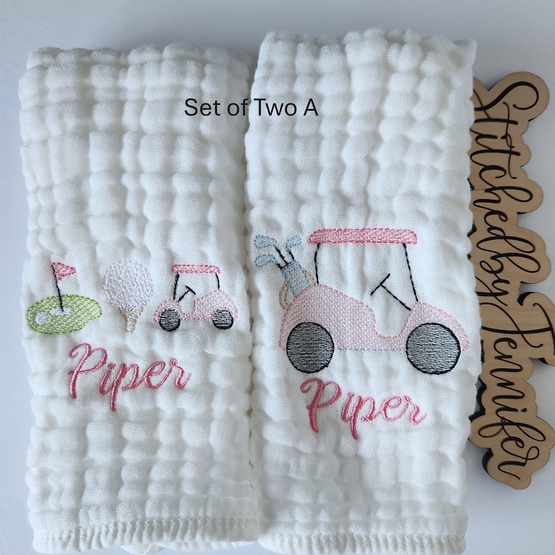 May include: Set of two white muslin burp cloths with pink and green embroidered golf cart designs and the name "Piper". The text "Set of Two A" is visible in the top right corner.