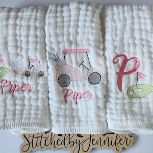 May include: Three white muslin burp cloths with pink and green embroidery. The center cloth features a pink golf cart with the name "Piper" embroidered below. The left cloth has a golf ball, golf club, and green golf course with the name "Piper" embroidered below. The right cloth has a pink letter "P" and a green golf course.