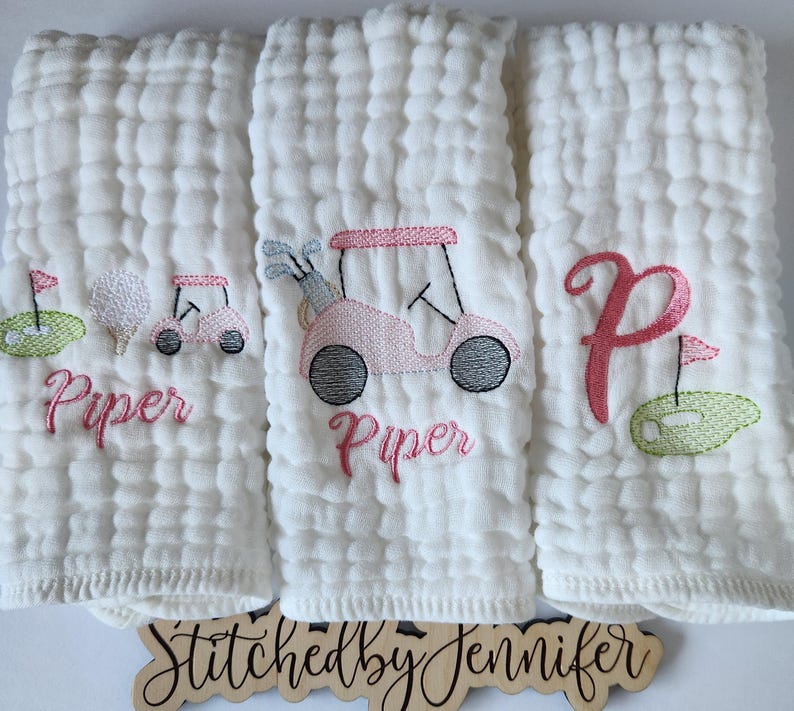 May include: Three white muslin burp cloths with pink and green embroidery. The center cloth features a pink golf cart with the name "Piper" embroidered below. The left cloth has a golf ball, golf club, and green golf course with the name "Piper" embroidered below. The right cloth has a pink letter "P" and a green golf course.
