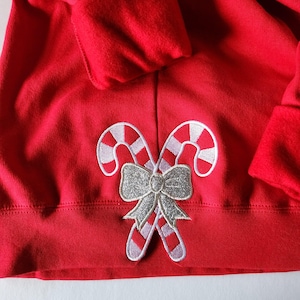 May include: Red sweatshirt with a festive candy cane and bow design. The candy canes are red and white, and the bow is silver. The sweatshirt is a solid red color.