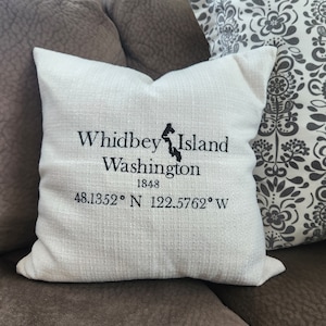 May include: White throw pillow with black embroidery that reads "Whidbey Island Washington 1848 48.1352° N 122.5762° W".