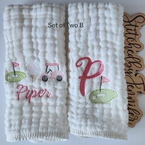 May include: Set of two white burp cloths with embroidered designs. One cloth features a pink golf cart, golf ball, and flag with the name "Piper" embroidered below. The other cloth features a pink letter "P" with a green golf course and flag. The text "Set of Two B" is visible at the top of the image.