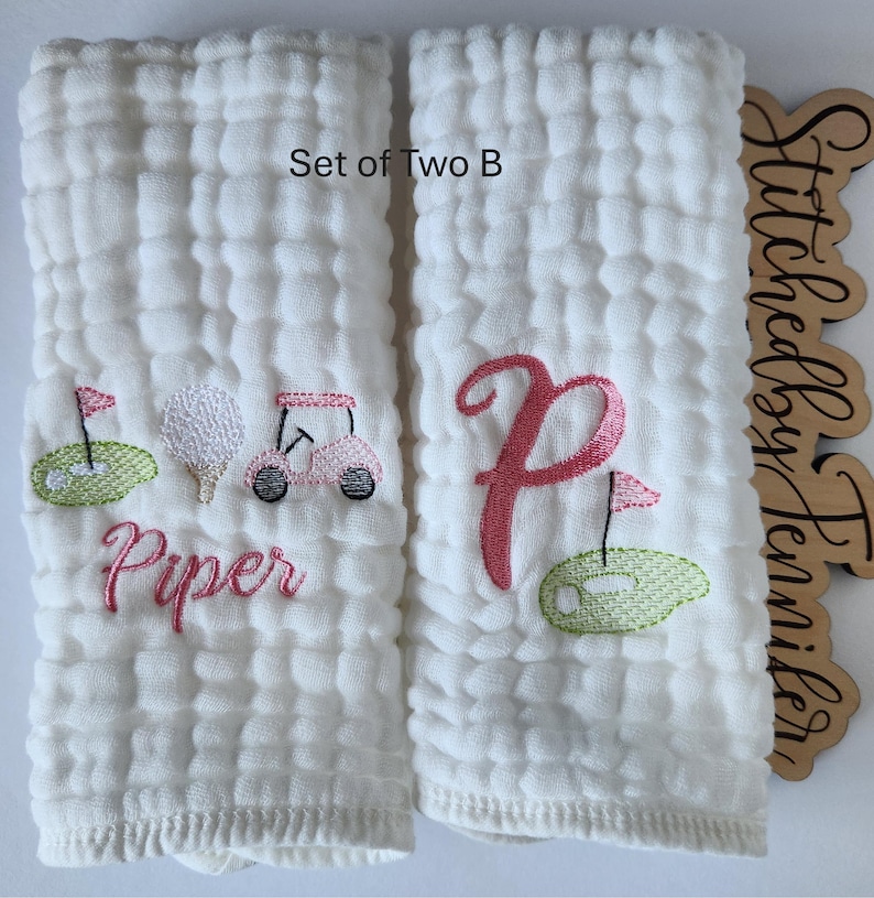 May include: Set of two white burp cloths with embroidered designs. One cloth features a pink golf cart, golf ball, and flag with the name "Piper" embroidered below. The other cloth features a pink letter "P" with a green golf course and flag. The text "Set of Two B" is visible at the top of the image.
