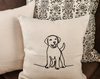 May include: Off-white square throw pillow with a black line drawing of a Labrador puppy. The pillow has a textured weave and is placed on a brown leather couch with other decorative pillows.