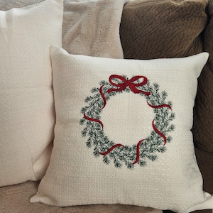 May include: A white throw pillow with an embroidered green wreath and a red ribbon bow.