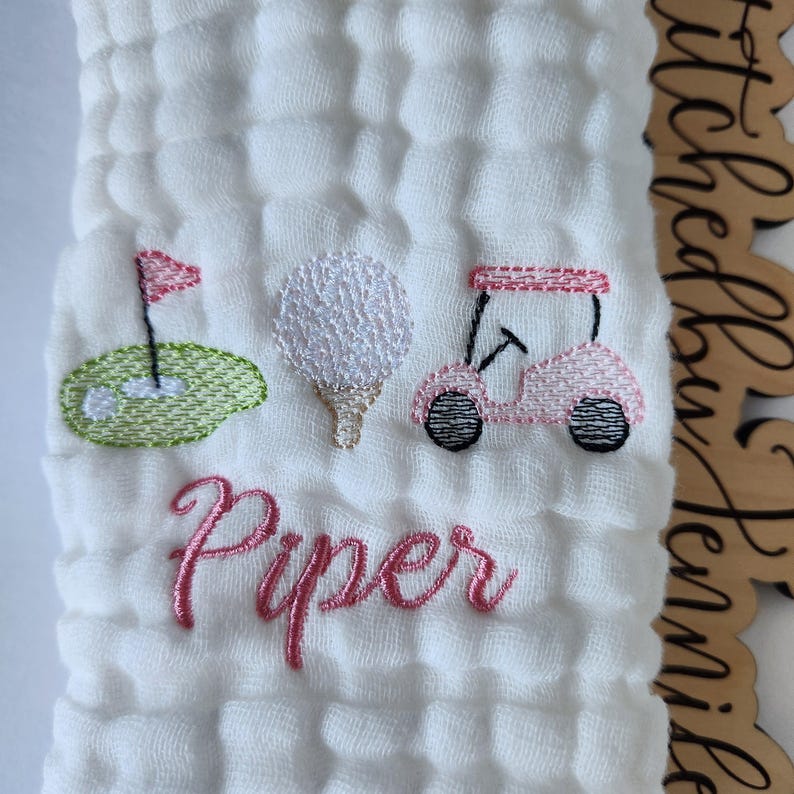 May include: White muslin baby burp cloth with pink embroidered golf cart, golf ball, and golf flag. The name "Piper" is embroidered in pink below the golf theme.