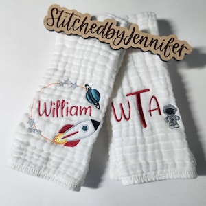 May include: Two white burp cloths with embroidered designs. One cloth features a rocket ship, a planet, stars, and the name "William". The other cloth features the letters "WTA" and an astronaut. Both cloths have a waffle-like texture.