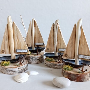 May include: Four miniature wooden sailboats with light brown sails and dark blue hulls. Each boat rests on a birch bark base with an anchor and small plants. The boats have labels that read "Hazy Days", "Castaway", "Liberty", and "Saltspray".