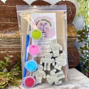 May include: A clear plastic bag contains a craft kit with robot-shaped plaster figures, paint pots in pink, blue, green, and red, and a paintbrush. A card with the text "Travel Imagination" and "Live simply" is also included.