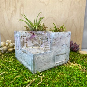 May include: A concrete planter with a unique architectural design, featuring a small staircase and a textured surface. The planter is filled with green plants and is sitting on a bed of moss.