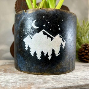 May include: A cylindrical black planter with a white mountain and forest design, a crescent moon, and stars. The planter has a textured finish with gold and blue accents. The planter is holding a green plant.