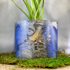 May include: A blue, silver, and gold marbled concrete planter with a green plant growing inside. The planter has a smooth, polished finish.