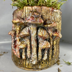 May include: A gold and red ceramic planter shaped like a tree stump with mushrooms growing out of it. The planter has a rustic, textured surface.