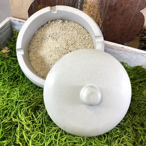May include: A gray concrete ashtray with a lid. The ashtray is filled with gray sand.