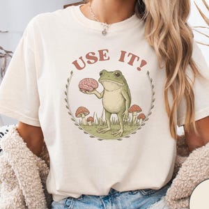 May include: Cream-colored t-shirt featuring a cartoon frog holding a brain, surrounded by mushrooms, with the text "USE IT!". The shirt also has the text "AUTHENTIC COMFORT COLORS 100% COTTON".