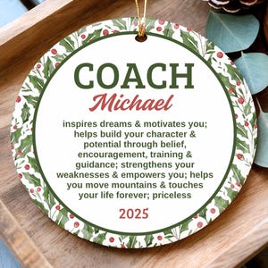 May include: A round ceramic ornament with a glazed finish, featuring a green holly and red berry border. The text reads: COACH Michael, inspires dreams & motivates you; helps build your character & potential through belief, encouragement, training & guidance; strengthens your weaknesses & empowers you; helps you move mountains & touches your life forever; priceless, 2025.