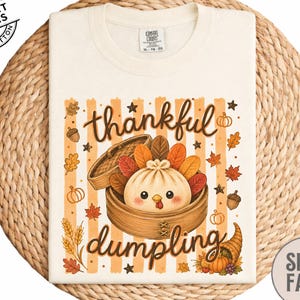May include: A cream-colored t-shirt featuring a graphic of a dumpling inside a bamboo steamer with turkey feathers. The text reads "thankful dumpling" in brown script. The design includes autumn leaves, acorns, and pumpkins on a striped background.
