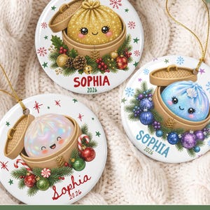May include: Three round white ceramic ornaments featuring illustrations of cute dumplings in bamboo steamers. Each ornament is decorated with pine branches, ornaments, and the name Sophia with the year 2026. Gold strings are attached for hanging.