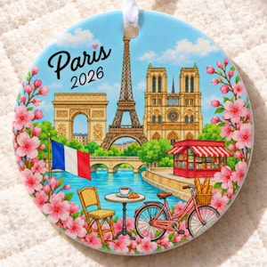 May include: A round ceramic ornament featuring a colorful illustration of Paris landmarks including the Eiffel Tower, Arc de Triomphe, and Notre Dame. The scene includes a French flag, a bicycle, a cafe table, and pink cherry blossoms with the text Paris 2026.