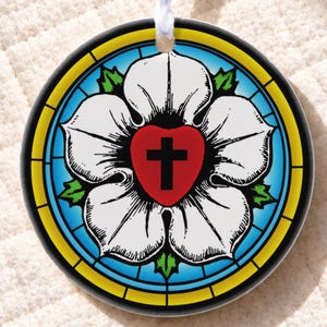 May include: A circular ceramic ornament featuring a Luther Rose design with a white five-petaled flower, a central red heart containing a black cross, and a blue background framed by a yellow border, suspended by a white ribbon.