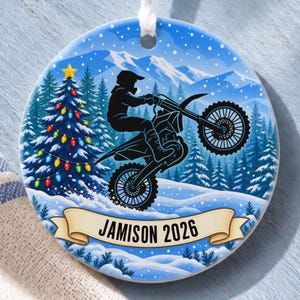 May include: A round ceramic Christmas ornament featuring a silhouette of a person riding a dirt bike in a snowy mountain landscape with a decorated pine tree. A beige banner at the bottom displays the text JAMISON 2026 in black font.