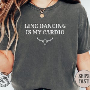 Puede incluir: Una camiseta gris oscura con el texto "Line Dancing is My Cardio" en blanco. Un contorno blanco de una cabeza de toro está debajo del texto.