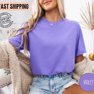 May include: A violet-colored t-shirt with short sleeves. The shirt is made of 100% cotton and has the words "Authentic Comfort Colors" printed on it. The words "Fast Shipping" are also visible.