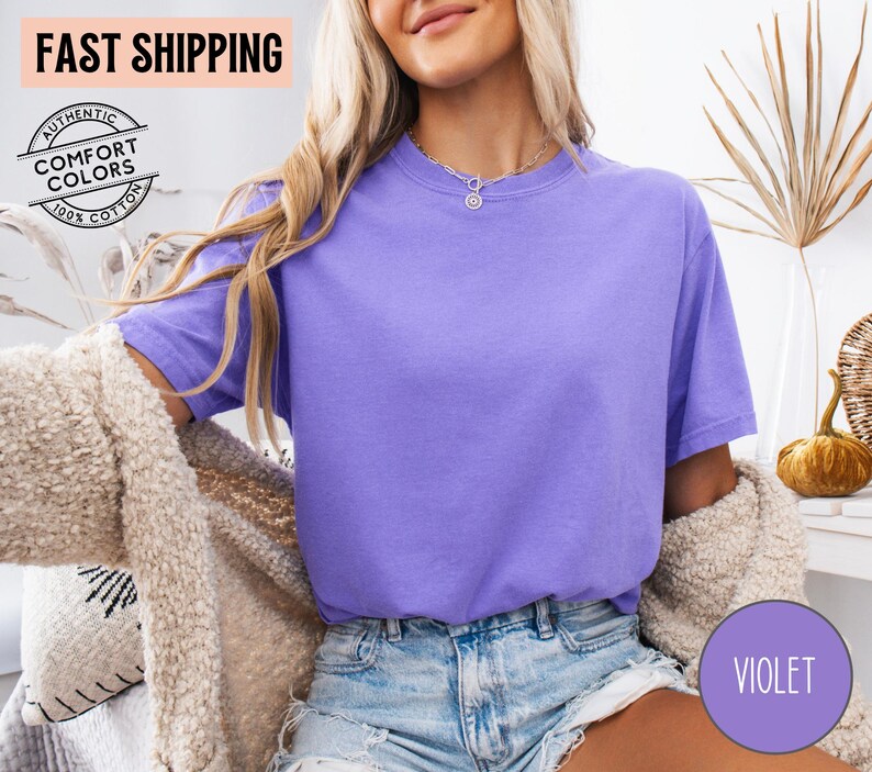 May include: A violet-colored t-shirt with short sleeves. The shirt is made of 100% cotton and has the words "Authentic Comfort Colors" printed on it. The words "Fast Shipping" are also visible.