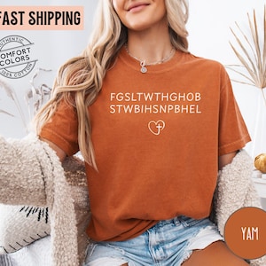 May include: A burnt orange t-shirt with white text and a heart with a cross graphic. The shirt has the words "AUTHENTIC COMFORT COLORS 100% COTTON" printed on it. The shirt is made of a soft material.